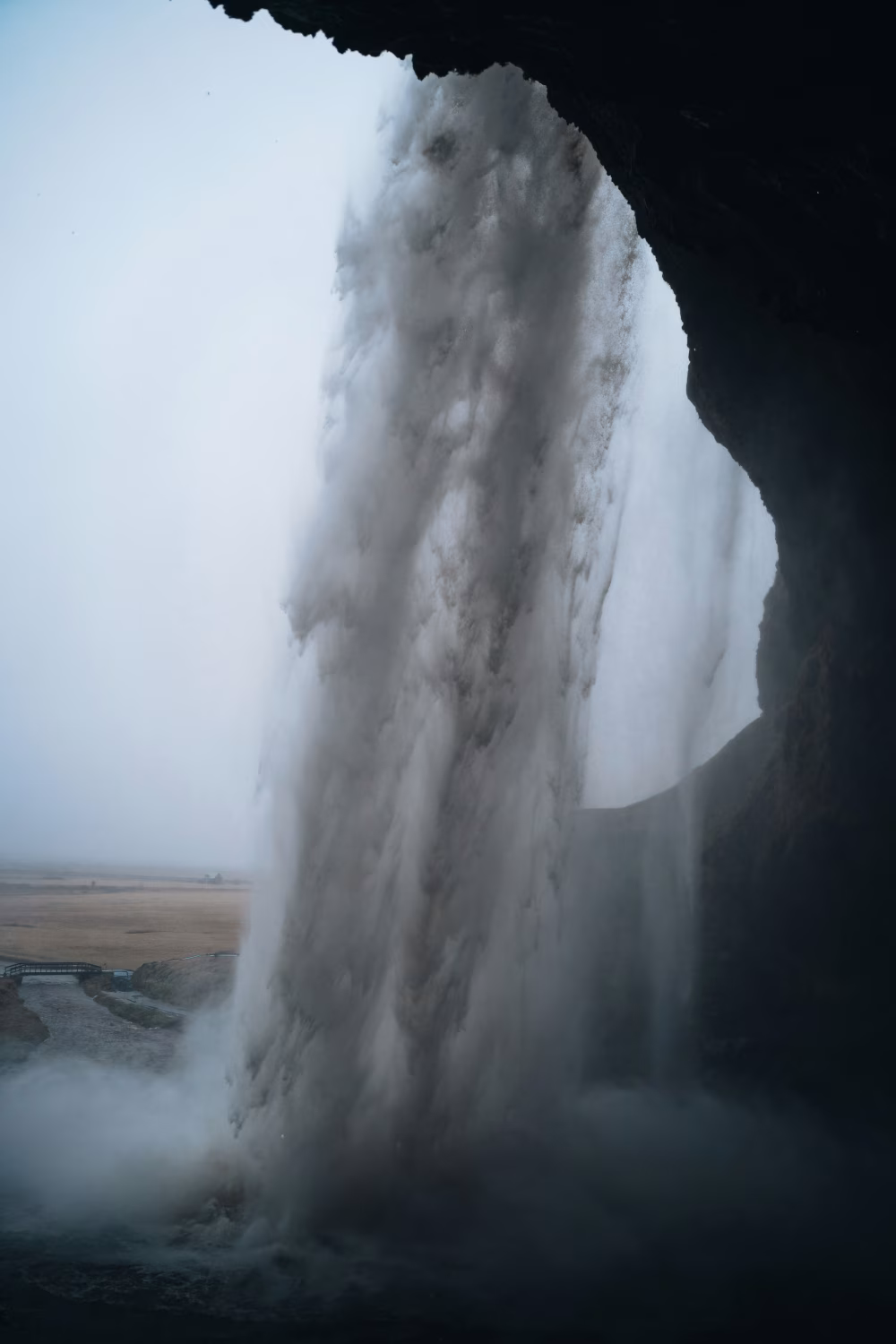 Dramatic Waterfall Seen From Behind a Cave With Misty Spray and Moody Landscape View