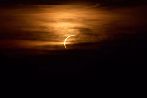 Partial Solar Eclipse Crescent Sun Behind Orange Clouds Over Dark Silhouette Landscape At Dusk