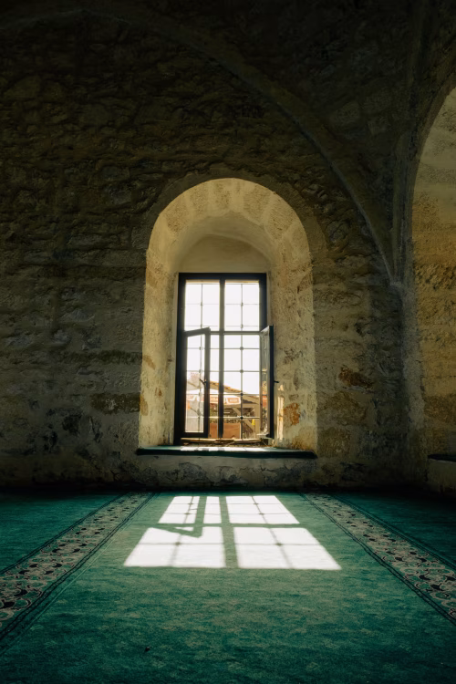 Sunlit Stone Arch Window Casting Shadow On Green Prayer Carpet Inside Historic Mosque Interior